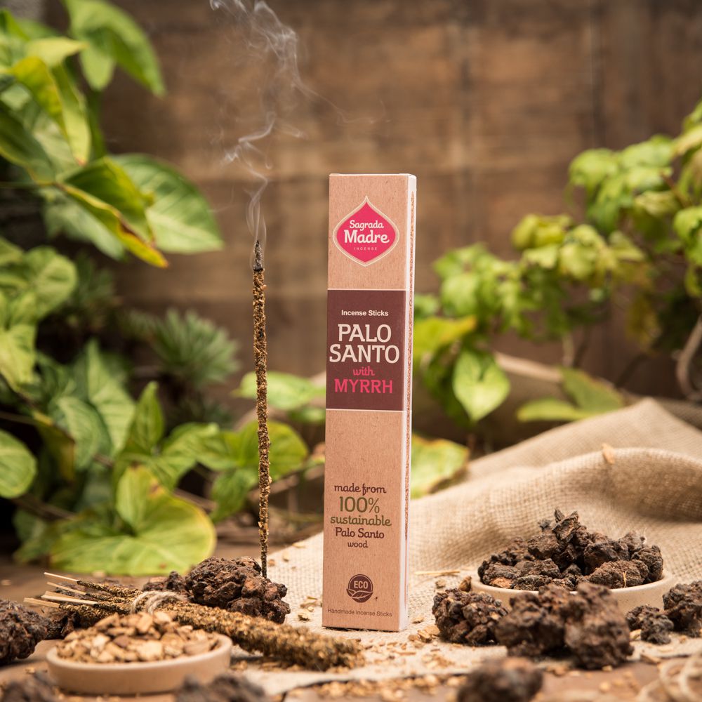 A box of Sagrada Madre Palo Santo incense sticks with myrrh, surrounded by natural elements and incense smoke rising in the background.